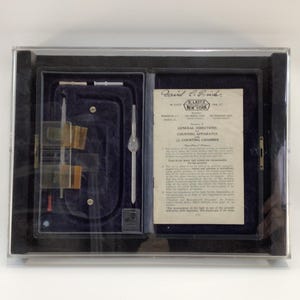 May include: A vintage scientific instrument set in a black case with a clear lid. The set includes glass slides, a pipette, and a printed document with the text "David C. Erich" and "E. Leitz New York". The interior is lined with dark blue velvet.