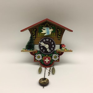 May include: A wooden cuckoo clock with a red roof, a white bird figure, and a dark circular clock face with white numbers. The base features painted white flowers, a red shield with a white cross, a green pine tree, a mushroom, and hanging weights.