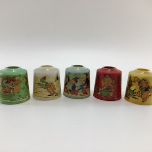 May include: Five vintage thimble holders in various colors, each featuring a different cartoon character illustration. The thimbles are green, off-white, blue, red, and yellow. The illustrations include a cat, a turtle, Mickey Mouse, an elephant, and Winnie the Pooh.