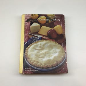 May include: A hardcover cookbook titled The Illustrated Encyclopedia of American Cooking by Editors of Favorite Recipes Press. The cover features a photograph of a baked pie, various cheeses, and fresh fruit on a wooden cutting board surface.