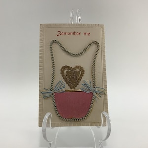May include: A vintage Valentine's Day card with a pink heart-shaped pouch and the text "Remember me". The pouch is decorated with gold rope and blue and white ribbon bows.