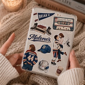 Puede incluir: Funda transparente para teléfono decorada con pegatinas. Las pegatinas incluyen una camiseta de hockey azul y blanca con el nombre "Graham" y el número "44", una cinta de cassette, una gorra de béisbol y una pareja abrazándose.