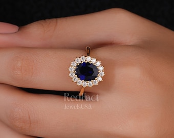Könnte beinhalten: Ein goldener Ring mit einem großen, ovalen, dunkelblauen Edelstein, der von kleineren, klaren Steinen umgeben ist. Der Ring hat ein dünnes Goldband und eine aufwendige Fassung. Der Ring wird auf einer Hand vor dunklem Hintergrund gezeigt.