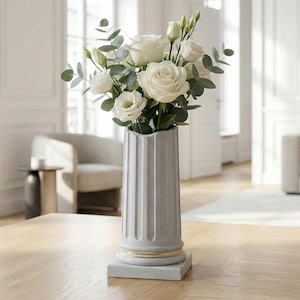 Op de afbeelding: Een grijze, gegroefde vaas in de vorm van een klassieke zuil met een boeket witte rozen en groen. De vaas heeft een gouden band aan de basis en staat op een vierkante basis. De opstelling staat op een houten tafel.