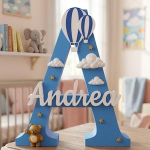 Op de afbeelding: Een blauwe houten letter "A" met de naam "Andrea" in witte letters. De letter is versierd met witte wolken, gouden sterren en twee blauw-witte luchtballonnen. Er is ook een kleine teddybeer aanwezig.