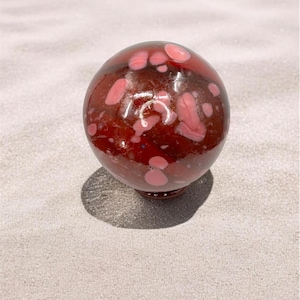May include: A decorative glass sphere with a deep red base color and pinkish-white spots. The sphere sits on a small, round base. The background is a light gray surface.