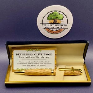 May include: A wooden pen and a pen holder are displayed in a black box with a gold trim. The pen is made of Bethlehem olive wood and has gold accents. A card inside the box reads "BETHLEHEM OLIVE WOOD From Bethlehem, The Holy Land." A round logo is above the box.