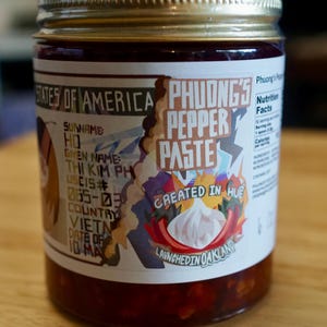 May include: A glass jar of Phuong's Pepper Paste with a gold lid. The label features text and graphics, including the words "Pepper Paste" and "Created in Hue, Launched in Oakland." The jar is filled with a red paste.