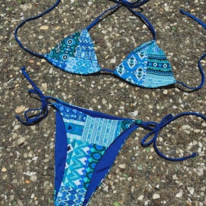 May include: A two-piece bikini set in shades of blue. The top is a triangle style with tie straps, and the bottoms are a classic cut. Both pieces feature a patchwork design with geometric and abstract patterns.