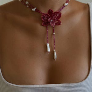May include: A magenta beaded necklace featuring a flower pendant and dangling pearl accents. The necklace has a delicate design with small beads and larger pearls, creating an elegant and feminine look. The necklace is a statement piece.