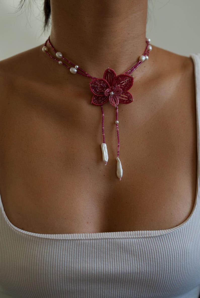 May include: A magenta beaded necklace featuring a flower pendant and dangling pearl accents. The necklace has a delicate design with small beads and larger pearls, creating an elegant and feminine look. The necklace is a statement piece.