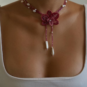 May include: A handmade necklace featuring a flower pendant and pearl accents. The necklace has a deep pink beaded design with a flower-shaped pendant. Two strands of pearls and two dangling white baroque pearls complete the design.