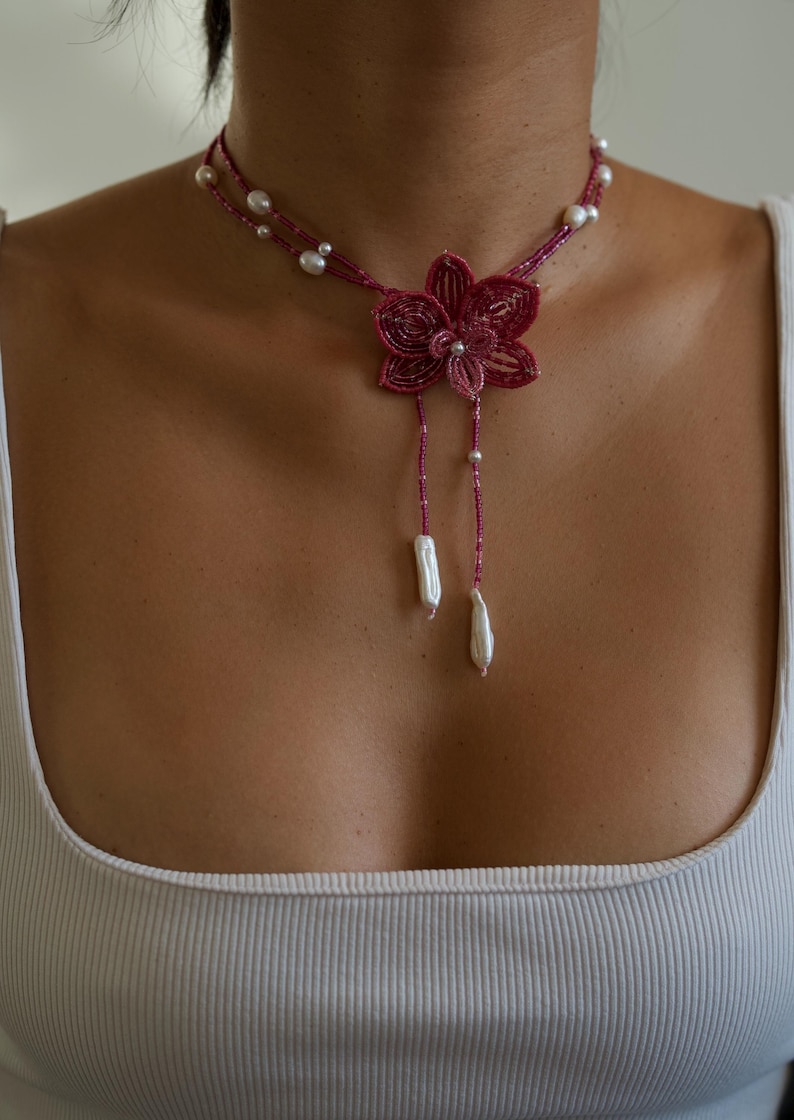 May include: A handmade necklace featuring a flower pendant and pearl accents. The necklace has a deep pink beaded design with a flower-shaped pendant. Two strands of pearls and two dangling white baroque pearls complete the design.