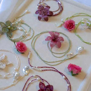May include: An assortment of handmade jewelry pieces, including beaded necklaces and bracelets, featuring floral designs and freshwater pearls. The color palette includes shades of green, white, and burgundy, with real pink roses scattered throughout.