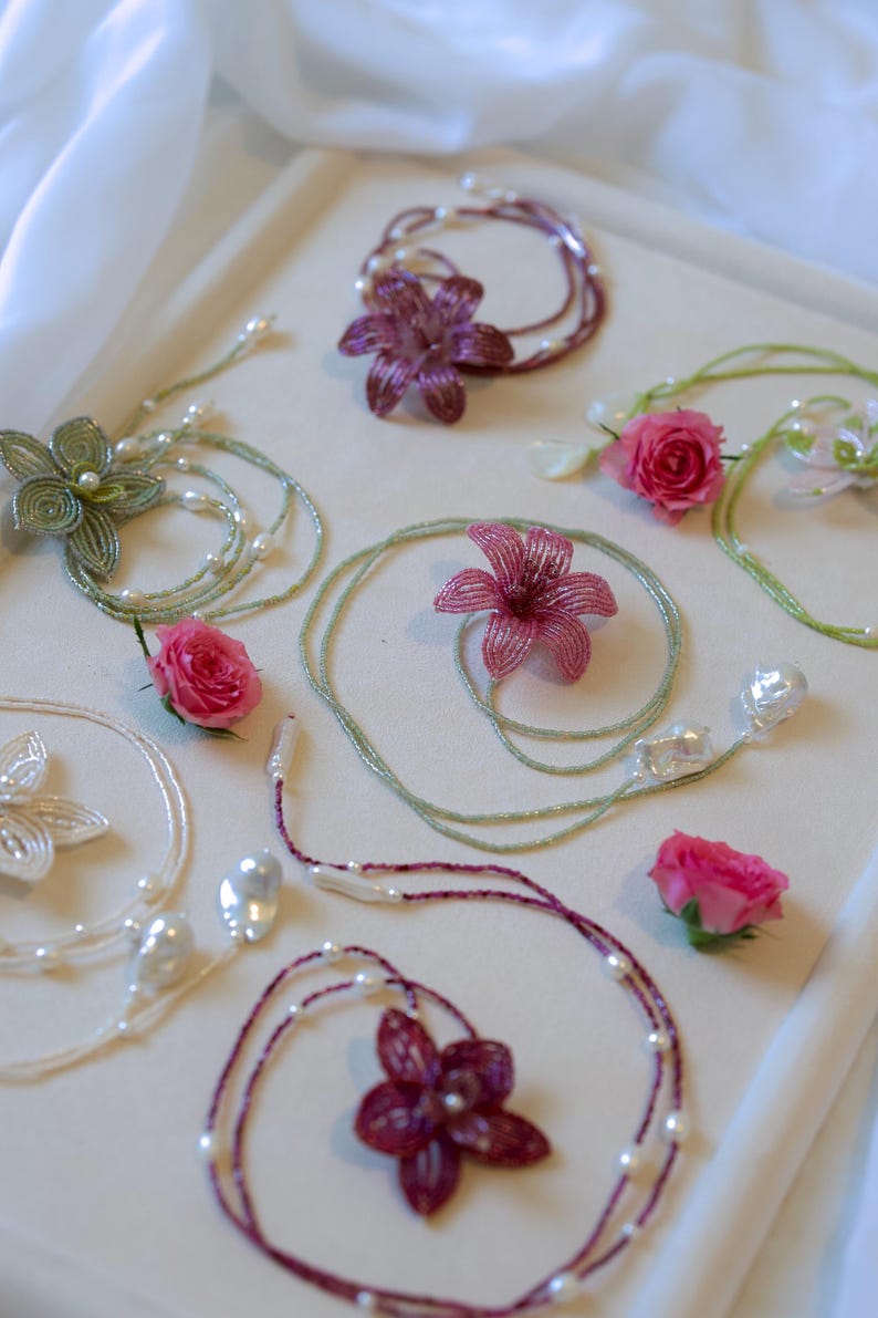 May include: An assortment of handmade jewelry pieces, including beaded necklaces and bracelets, featuring floral designs and freshwater pearls. The color palette includes shades of green, white, and burgundy, with real pink roses scattered throughout.