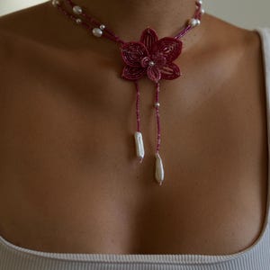 May include: A handcrafted necklace featuring a burgundy beaded flower pendant with dangling white pearl accents. The necklace has a double strand design with small pearls and a delicate, feminine aesthetic.