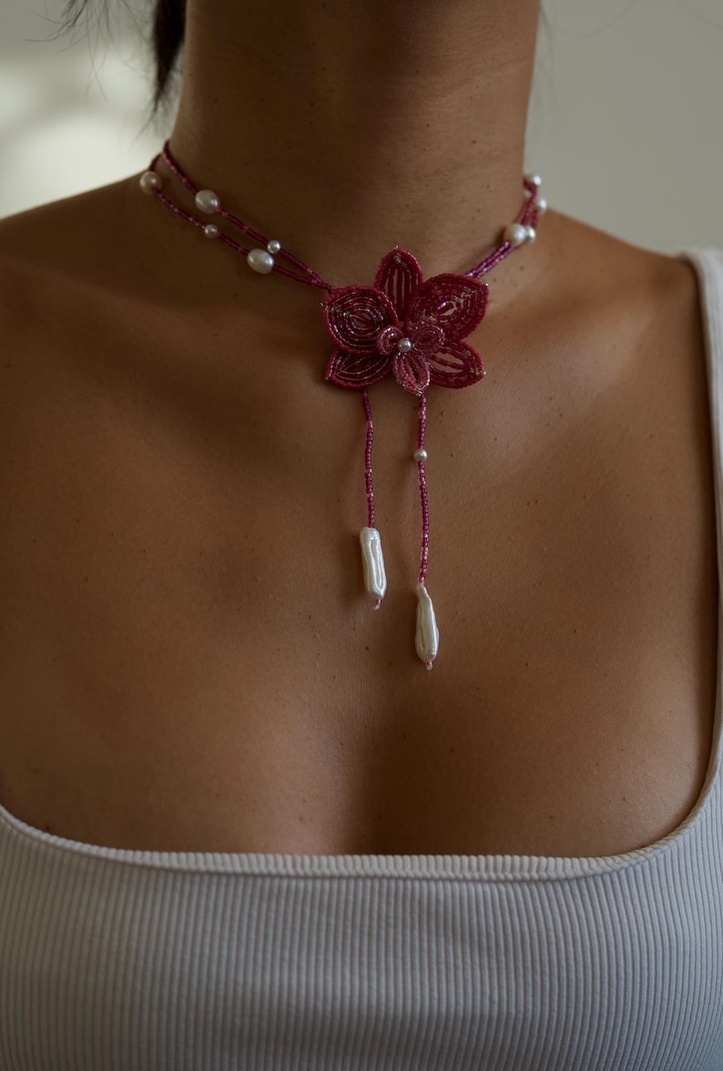 May include: A handcrafted necklace featuring a burgundy beaded flower pendant with dangling white pearl accents. The necklace has a double strand design with small pearls and a delicate, feminine aesthetic.