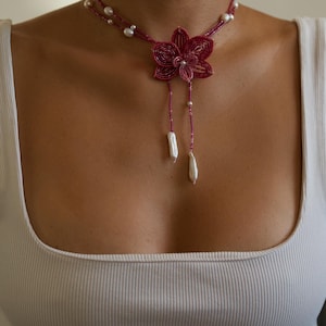 May include: A handmade necklace featuring a burgundy flower pendant with dangling white pearls. The necklace has a double strand design with small white pearls and a delicate beaded chain. A unique floral jewelry piece.