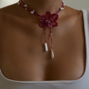 May include: A beaded choker necklace featuring a magenta flower pendant with dangling white pearl accents. The necklace has a double strand design with small white pearls. The flower pendant is the focal point of the necklace.