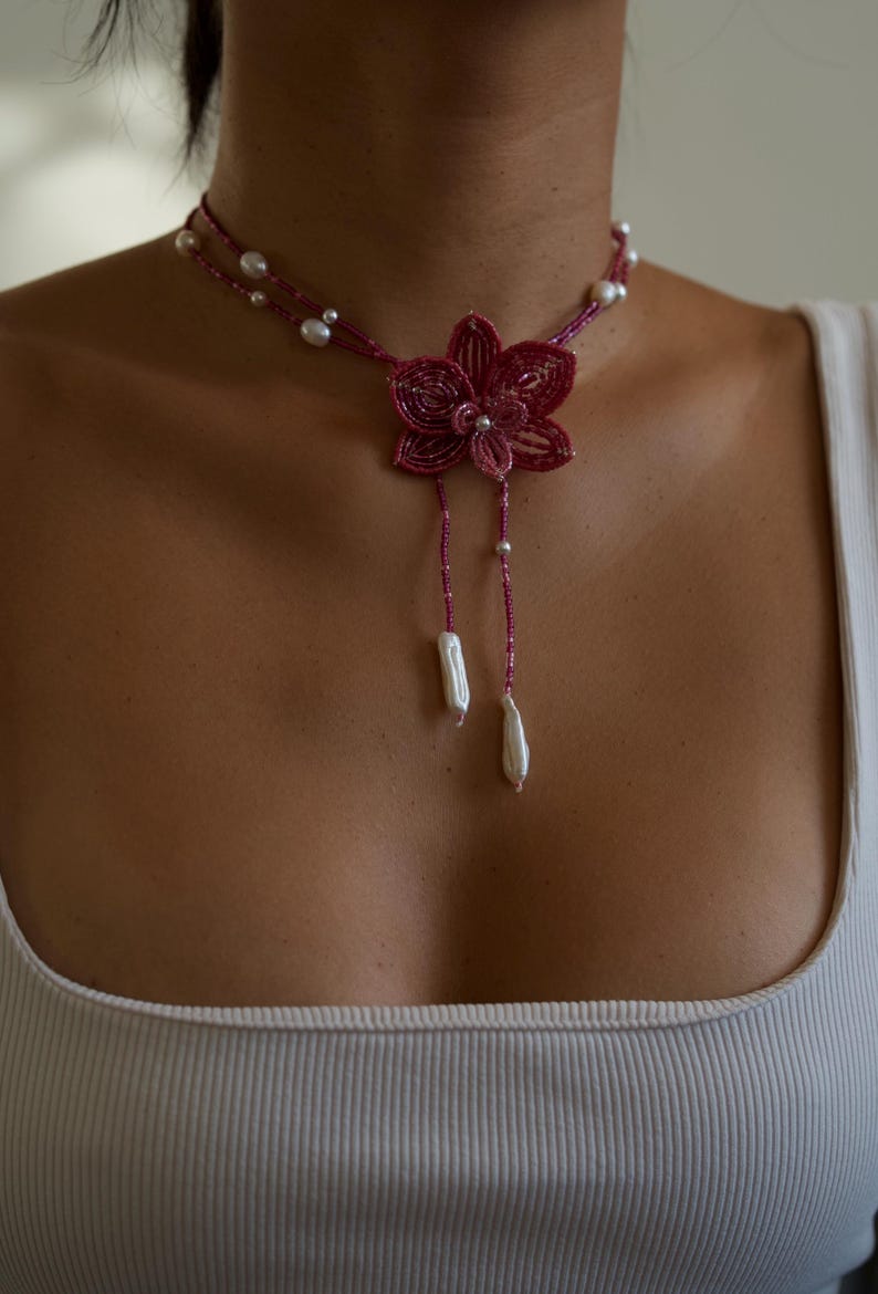 May include: A beaded choker necklace featuring a magenta flower pendant with dangling white pearl accents. The necklace has a double strand design with small white pearls. The flower pendant is the focal point of the necklace.