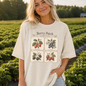 May include: A cream-colored t-shirt featuring a graphic print titled Berry Patch Collection. The design includes four rectangular labels with botanical illustrations of strawberries, blueberries, blackberries, and raspberries with text.
