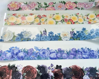 May include: Floral washi tape rolls featuring various flower designs. The rolls display a variety of blooms in shades of pink, yellow, blue, and burgundy. The tape is transparent, allowing the floral patterns to be visible.