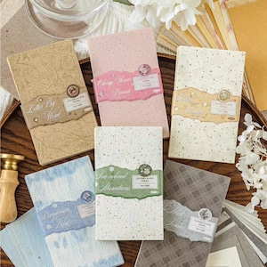 May include: A selection of decorative paper notebooks with different designs and text, including "Letter By Wood", "Cherry Flower Dream", "Daydream Blue", and "Tea-colored Mountains". The notebooks are in shades of beige, pink, blue, and grey.