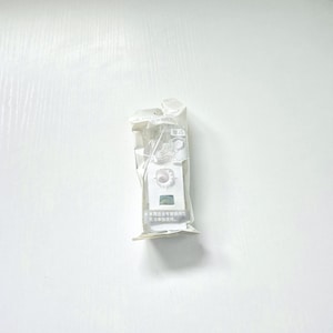 Peut inclure: Un petit objet rectangulaire blanc est emballé dans un emballage plastique transparent. L'objet présente un motif circulaire avec une base carrée. L'emballage porte du texte japonais et est scellé en haut.