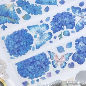 May include: Decorative washi tape featuring blue butterflies and hydrangea flowers. The tape has a transparent background and is adorned with watercolor-style illustrations. The design includes various shades of blue and hints of pink.