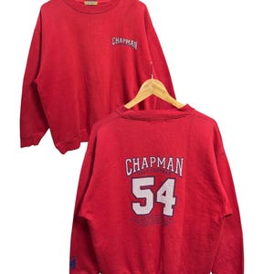 May include: Red Chapman sweatshirt with the word "Chapman" printed on the front and back. The back of the sweatshirt also features the number "54". The sweatshirt is hanging on a wooden hanger.