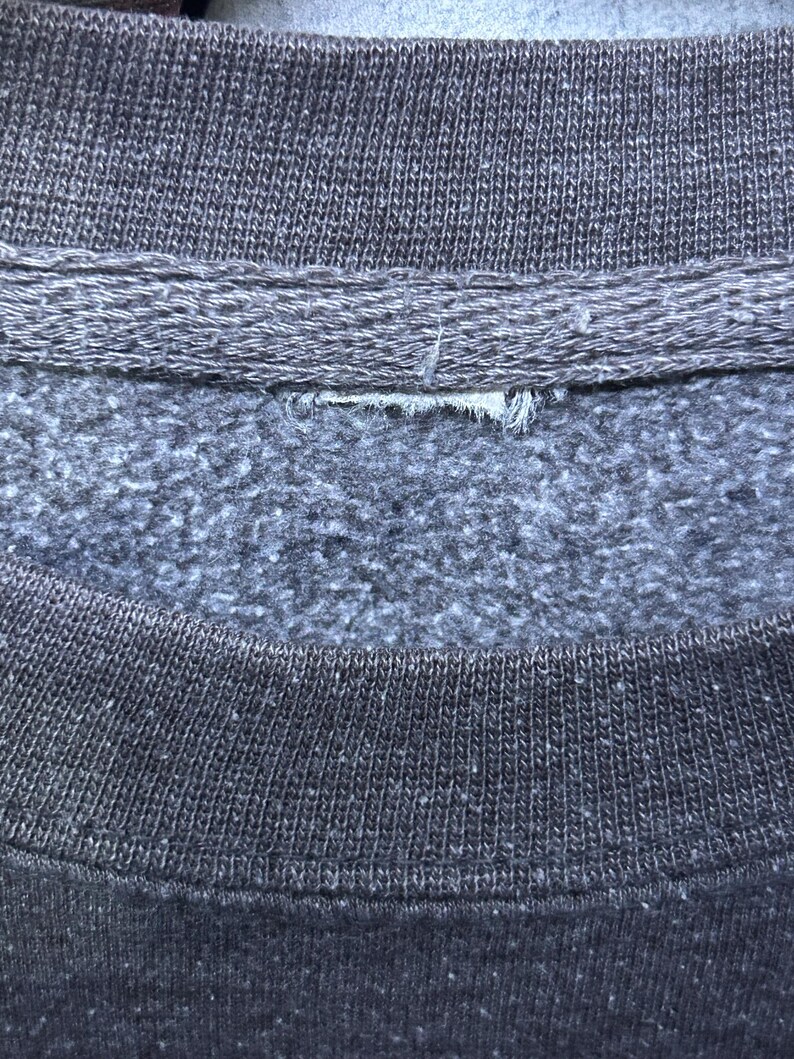 May include: Close-up of a gray sweatshirt collar. The collar is a darker gray than the body of the sweatshirt. The fabric has a textured appearance. The image shows the neckline and part of the shoulder area of the garment.