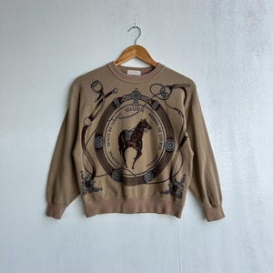 Op de afbeelding: Tan sweatshirt met een paard en een ontwerp met een paardenthema. De afbeelding toont een bruin paard in een cirkelvormige lijst met tekst en decoratieve elementen. De sweatshirt heeft lange mouwen en een ronde hals.