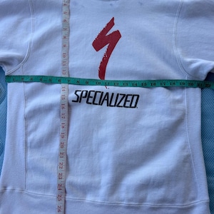 May include: White sweatshirt with a red Specialized logo and text. The sweatshirt has a crew neck and long sleeves. The chest measures approximately 20 inches across. The garment is on a wooden hanger.