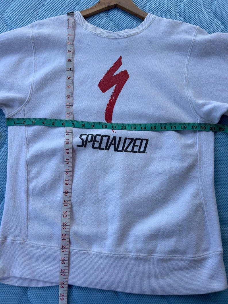 May include: White sweatshirt with a red Specialized logo and text. The sweatshirt has a crew neck and long sleeves. The chest measures approximately 20 inches across. The garment is on a wooden hanger.