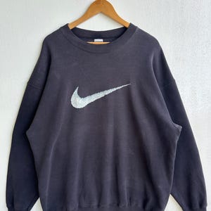 May include: A dark grey crew neck sweatshirt featuring a large Nike swoosh logo in white and grey. The sweatshirt is suspended on a wooden hanger. The fabric has a slightly faded appearance against a white backdrop.