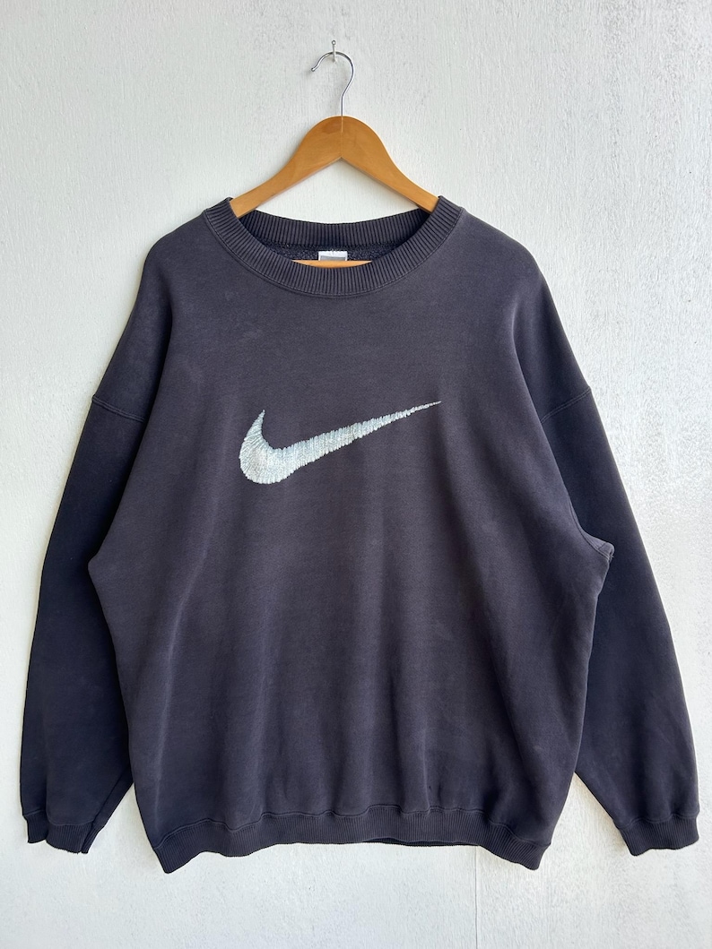 May include: A dark grey crew neck sweatshirt featuring a large Nike swoosh logo in white and grey. The sweatshirt is suspended on a wooden hanger. The fabric has a slightly faded appearance against a white backdrop.