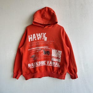 May include: An orange hooded sweatshirt with the word "HAWK" printed in white across the chest. The sweatshirt also features the words "BACKSIDE VARIAL" and other text. The design includes graphic elements. The garment is made of a soft material.