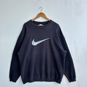 May include: A black crew neck sweatshirt featuring a large, silver Nike swoosh logo. The sweatshirt is suspended from a wooden hanger against a plain white backdrop. The design and long sleeves suggest a vintage or retro style, typical of sportswear.