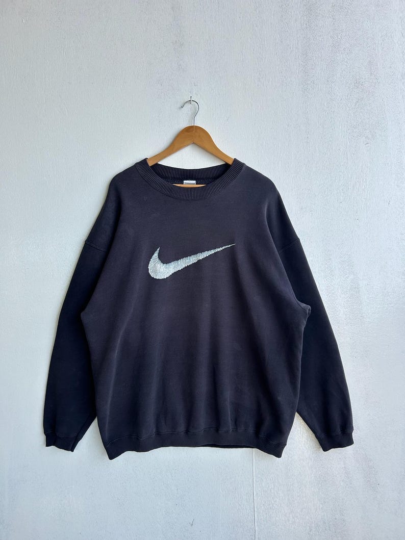 May include: A black crew neck sweatshirt featuring a large, silver Nike swoosh logo. The sweatshirt is suspended from a wooden hanger against a plain white backdrop. The design and long sleeves suggest a vintage or retro style, typical of sportswear.