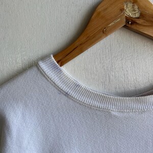 May include: Close-up of a white sweatshirt with a ribbed crew neck. The sweatshirt is hanging on a wooden hanger. The fabric appears soft and slightly textured.