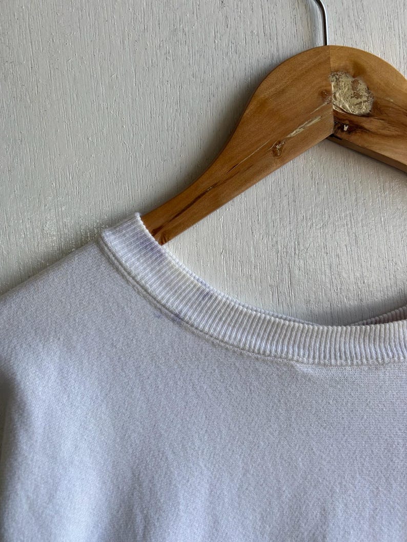 May include: Close-up of a white sweatshirt with a ribbed crew neck. The sweatshirt is hanging on a wooden hanger. The fabric appears soft and slightly textured.