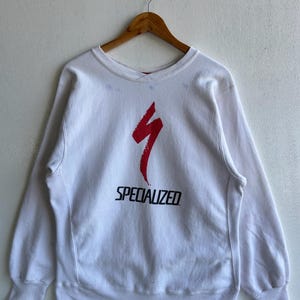 May include: White crew neck sweatshirt with the word "SPECIALIZED" in black block letters below a red stylized lightning bolt design. The sweatshirt is hanging on a wooden hanger against a white wall.