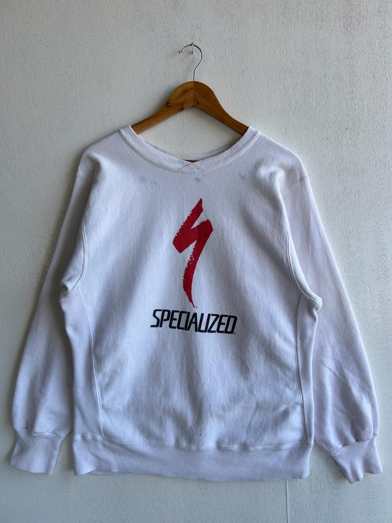 May include: White crew neck sweatshirt with the word "SPECIALIZED" in black block letters below a red stylized lightning bolt design. The sweatshirt is hanging on a wooden hanger against a white wall.