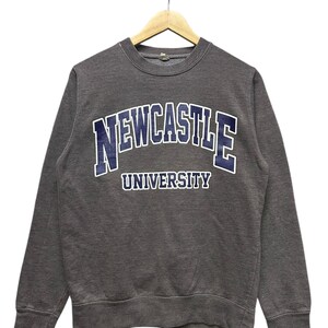 May include: A gray sweatshirt with the text "NEWCASTLE UNIVERSITY" in navy blue and white lettering. The crew neck sweatshirt is hanging on a wooden hanger against a white background. The long-sleeved top is made of a soft material.