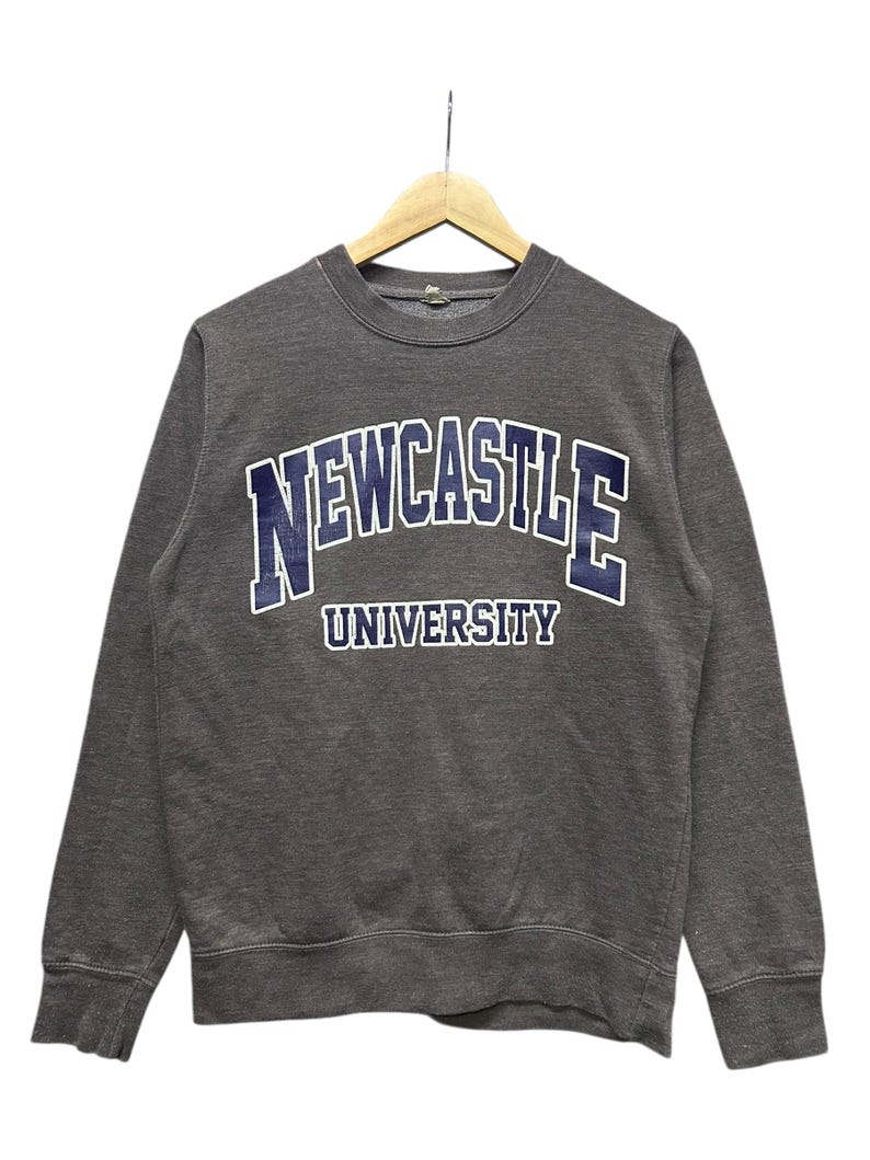 May include: A gray sweatshirt with the text "NEWCASTLE UNIVERSITY" in navy blue and white lettering. The crew neck sweatshirt is hanging on a wooden hanger against a white background. The long-sleeved top is made of a soft material.