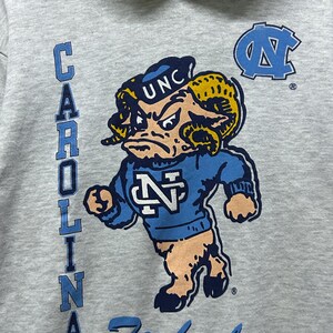 K&ouml;nnte beinhalten: Grauer Kapuzenpullover mit einem Cartoon-Widder-Maskottchen in einem blauen Pullover mit den Buchstaben "N" und "UNC". Das Wort "CAROLINA" ist vertikal auf der linken Seite aufgedruckt, und "Tarheels" steht unten in Blau.