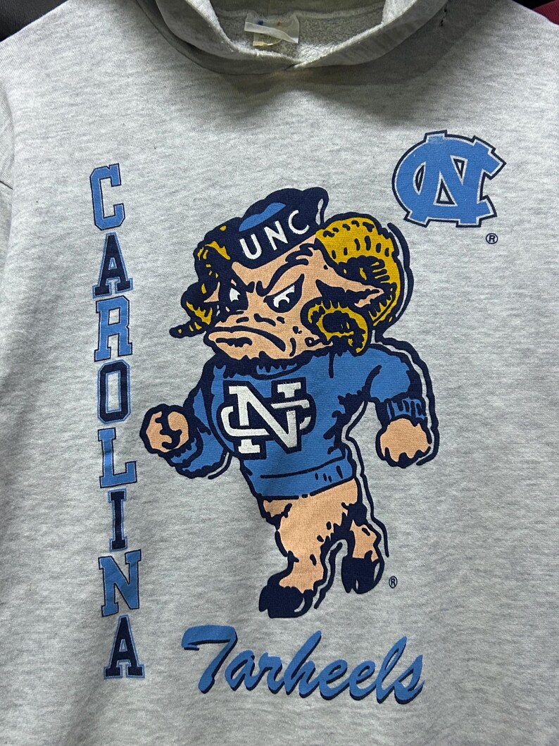 K&ouml;nnte beinhalten: Grauer Kapuzenpullover mit einem Cartoon-Widder-Maskottchen in einem blauen Pullover mit den Buchstaben "N" und "UNC". Das Wort "CAROLINA" ist vertikal auf der linken Seite aufgedruckt, und "Tarheels" steht unten in Blau.