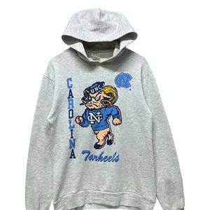 K&ouml;nnte beinhalten: Grauer Kapuzenpullover mit dem Logo der University of North Carolina Tar Heels. Das Design zeigt ein Cartoon-Widder-Maskottchen in Blau und Wei&szlig; mit den W&ouml;rtern "Carolina" und "Tar Heels" in Blau.