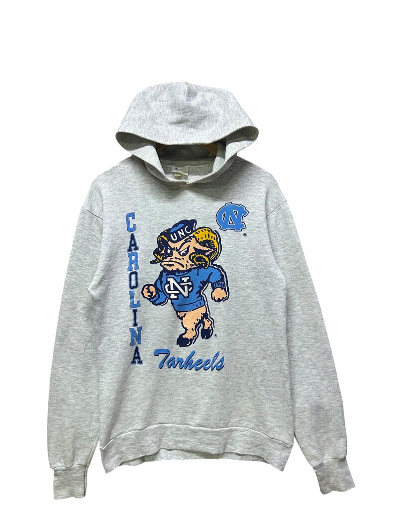 K&ouml;nnte beinhalten: Grauer Kapuzenpullover mit dem Logo der University of North Carolina Tar Heels. Das Design zeigt ein Cartoon-Widder-Maskottchen in Blau und Wei&szlig; mit den W&ouml;rtern "Carolina" und "Tar Heels" in Blau.