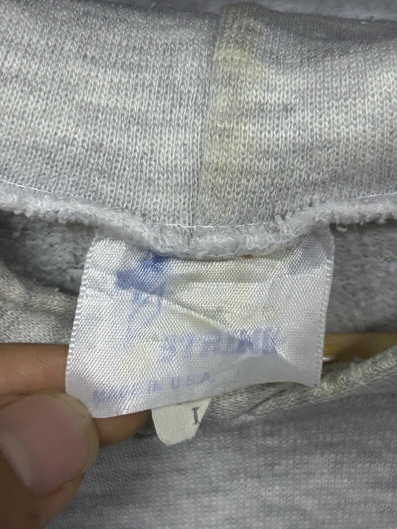 K&ouml;nnte beinhalten: Nahaufnahme eines grauen Kapuzenpullovers mit einem Etikett. Das Etikett ist wei&szlig; mit verblasstem blauem Text und den Worten "MADE IN USA". Der Kapuzenpullover ist aus einem weichen, strukturierten Stoff gefertigt.